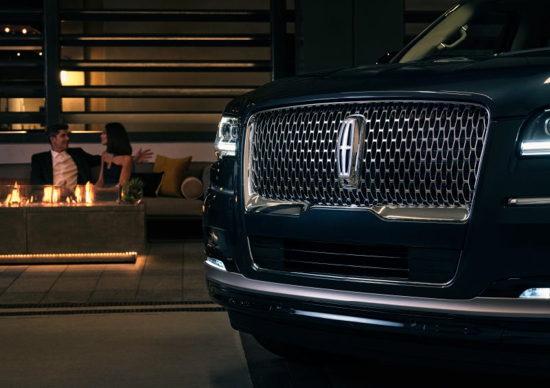 The signature Lincoln grille on the 2022 Lincoln Navigator SUV commands attention as a couple relaxes by a fire beyond | Stevens Creek Lincoln in San Jose CA