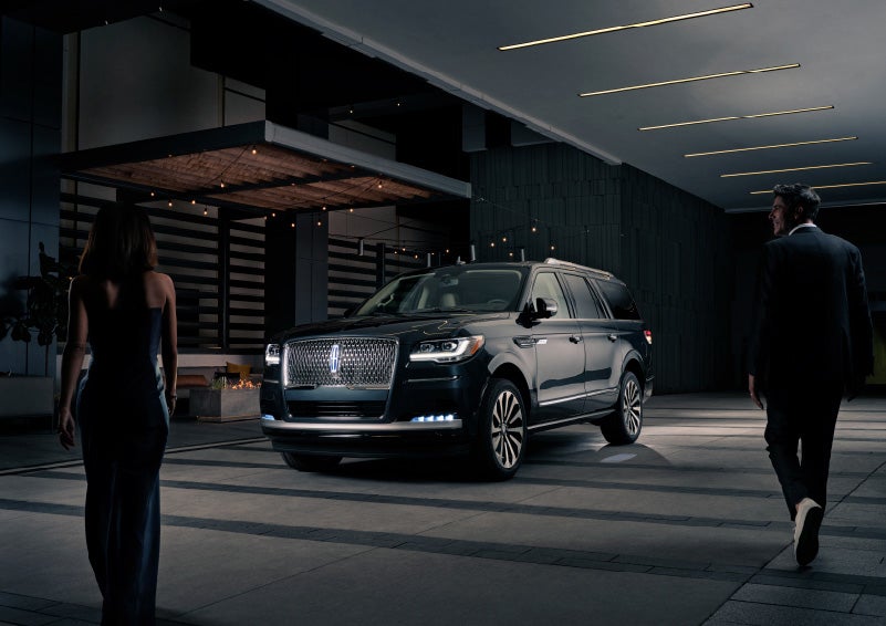 A man and woman in formal wear are approaching a 2022 Lincoln Navigator SUV at night as it lights with the Lincoln Embrace | Stevens Creek Lincoln in San Jose CA