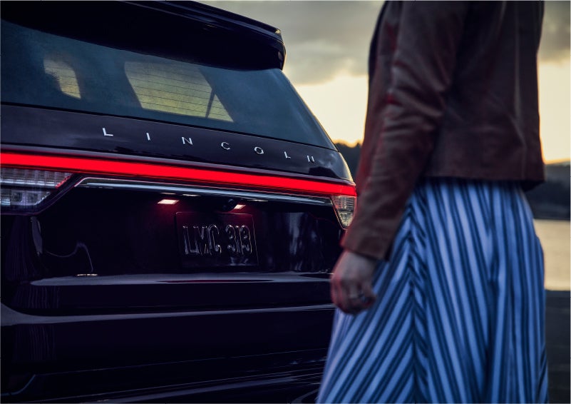A person is shown near the rear of a 2023 Lincoln Aviator® SUV as the Lincoln Embrace illuminates the rear lights | Stevens Creek Lincoln in San Jose CA