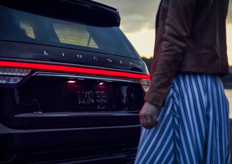 A person is shown near the rear of a 2024 Lincoln Aviator® SUV as the Lincoln Embrace illuminates the rear lights | Stevens Creek Lincoln in San Jose CA
