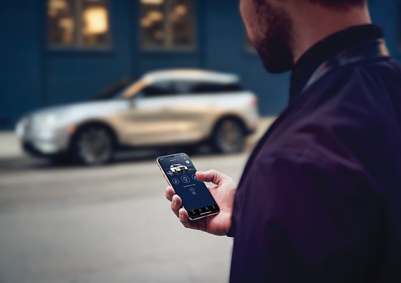 A person is shown interacting with a smartphone to connect to a Lincoln vehicle across the street. | Stevens Creek Lincoln in San Jose CA